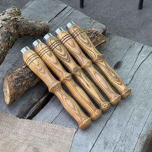 Könnte beinhalten: Fünf Werkzeuge mit Holzgriffen und Edelstahlaufsätzen sind diagonal angeordnet. Die Griffe haben ein hellbraunes Holzmaserungsmuster. Die Werkzeuge liegen auf einer verwitterten Holzoberfläche.