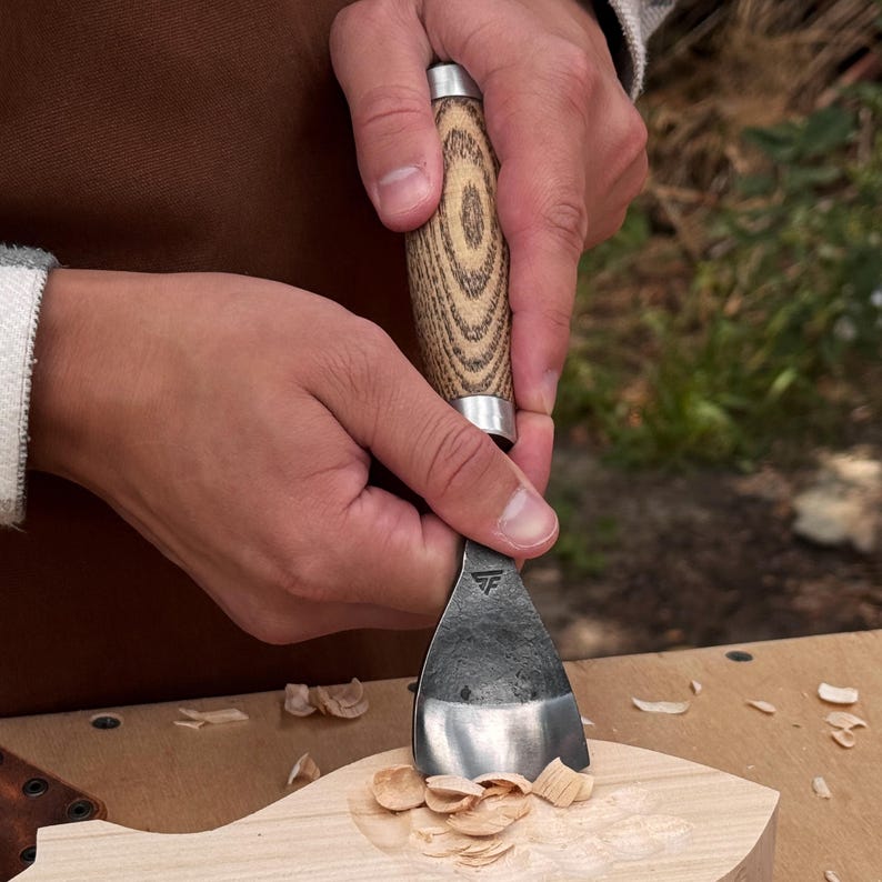 May include: A woodworking chisel with a textured wooden handle and a wide, curved metal blade featuring a small triangular logo. The tool is shown carving into a light-colored wooden block, creating small wood shavings on the surface.