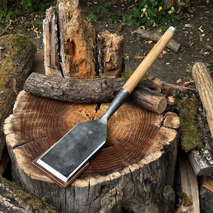 Op de afbeelding: Een handgereedschap met een breed, plat, donker metalen blad met een afgeschuinde rand en een lange, cilindrische houten steel. Het blad zit gedeeltelijk in een bruine lederen schede en het gereedschap ligt op een grote boomstronk.