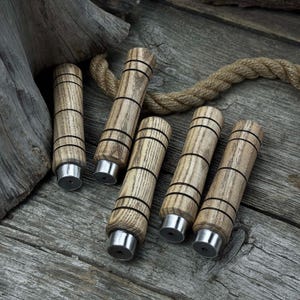 Może przedstawiać: Sześć drewnianych korków do butelek z jasnobrązowym wykończeniem i czarnymi paskami. Każdy korek ma srebrną metalową podstawę. Korki ułożone są na zwietrzałej drewnianej powierzchni z liną w tle.