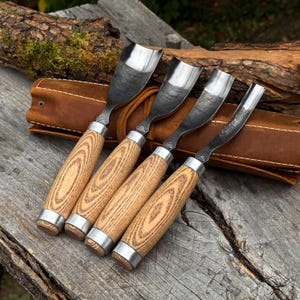 May include: A set of four wood carving gouges with curved metal blades and light brown wood grain handles. Each tool features a metal bolster and a small stamped logo near the base of the blade. The tools are arranged in a brown leather roll.