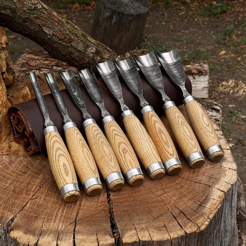 May include: A set of eight wood carving gouges with curved metal blades and light-coloured wooden handles featuring a prominent grain pattern. Each tool has a silver-coloured metal ferrule at the base of the handle, arranged in a row on a dark brown leather roll.