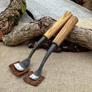 Pode incluir: Duas ferramentas manuais com lâminas curvas de metal cinza-escuro e cabos de madeira castanho-claro. Cada lâmina é protegida por uma pequena bainha quadrada de couro castanho fixada com quatro rebites de metal preto, com veios de madeira visíveis.