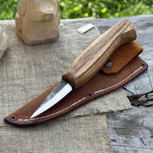 Wood carving knife with ash handle and leather sheath on workbench, top view showing compact forged blade for precise whittling.