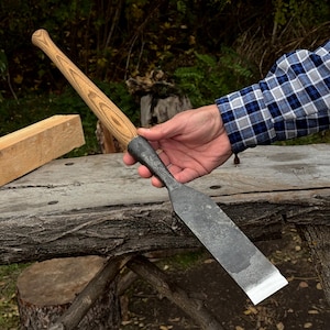 May include: A hand tool featuring a long, light-colored wooden handle with a visible grain pattern and a wide, flat, dark gray metal blade with a sharpened edge. The metal section connects to the handle with a tapered, textured dark gray bolster.