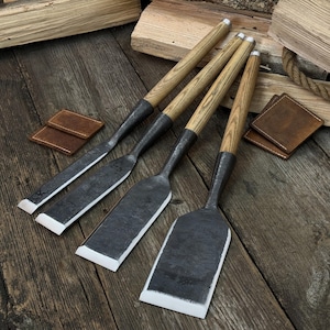 Puede incluir: Cuatro formones para carpintería con mangos de madera y hojas de metal están dispuestos sobre una superficie de madera. Los formones tienen un acabado de metal oscuro y mangos de madera de color claro. También se ven dos posavasos cuadrados de cuero.