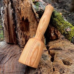 May include: A wooden mallet with a light brown finish. The mallet has a cylindrical handle and a rectangular head. The wood grain is visible. The mallet is resting on a weathered wooden surface.