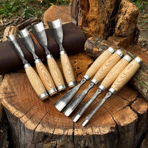 May include: A set of eight wood carving gouges with curved metal blades and light-coloured wooden handles. The tools feature silver-toned metal ferrules connecting the blades to the handles, with some gouges resting on a dark brown leather roll.