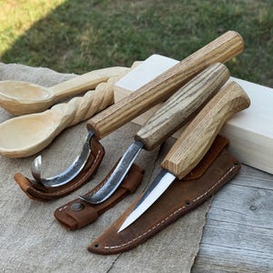 May include: A collection of wood carving tools, including a hook knife, a straight knife, and two wooden spoons. The knives have wooden handles and metal blades, with leather sheaths. The spoons are carved from light-colored wood.