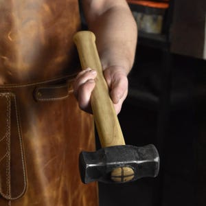 May include: A blacksmith's hammer with a wooden handle and a dark metal head. The hammer is held in a person's hand, with a leather apron visible in the background. The hammer head has a cross design.