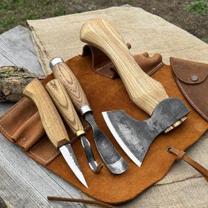 Puede incluir: Un conjunto de herramientas de carpintería, que incluye un hacha pequeña, cuchillos y cinceles, con mangos de madera y hojas de metal. Las herramientas se muestran sobre una superficie de cuero marrón, con un rollo y una bolsa de herramientas de cuero.
