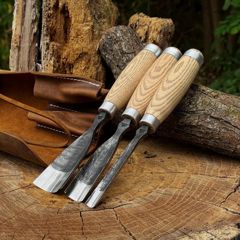 May include: Three wood carving chisels with light-coloured wooden handles and metal ferrules. The blades are forged steel with a dark, textured finish and varying widths, ranging from a wide gouge to a narrower blade, resting near a leather roll.