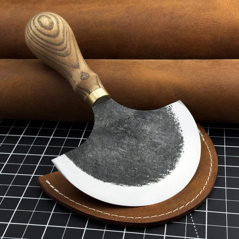 Pu&ograve; includere: Un coltello da lavoro in pelle con una lama curva grigio scuro e un manico in legno. Il manico presenta un motivo a venature chiare. Il coltello poggia su un fodero in pelle marrone, con uno sfondo a griglia in bianco e nero.