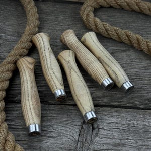 Peut inclure: Cinq manches de couteau en bois clair avec un motif de grain de bois et des viroles en métal argenté. Les manches sont disposés sur une surface en bois vieilli avec une longueur de corde en arrière-plan.