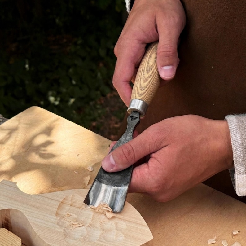 May include: A wood carving gouge with a curved metal blade and a light-colored wooden handle. The tool is being used to carve a shallow, rounded depression into a piece of light-colored wood, creating small wood shavings on the surface.