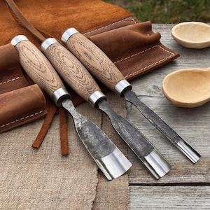 May include: Three wood carving chisels with wooden handles and metal blades are displayed on a wooden surface. A leather tool roll and two wooden spoons are also visible. The chisels have a rustic, handcrafted appearance.
