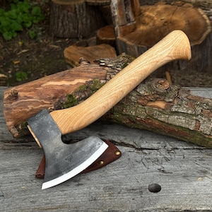 Puede incluir: Un hacha de mano con mango de madera clara curvada y cabeza de metal forjado oscuro con un filo pulido. Un pequeño logotipo geométrico estampado es visible en la hoja de metal, que descansa parcialmente dentro de una funda de cuero marrón.