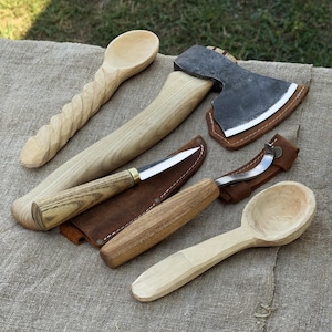 Puede incluir: Una colección de herramientas de madera hechas a mano, que incluye una cuchara con mango tallado, un hacha, un cuchillo en una funda de cuero y una cuchara curva. Las herramientas se exhiben sobre una superficie de lino de color neutro.