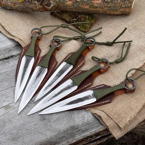K&ouml;nnte beinhalten: F&uuml;nf Wurfmesser mit silbernen Klingen und gr&uuml;n umwickelten Griffen werden gezeigt. Jedes Messer ist in einer braunen Lederh&uuml;lle verstaut. Gr&uuml;ne Kordeln sind an den Griffen befestigt. Die Messer sind auf einer Holzoberfl&auml;che angeordnet.