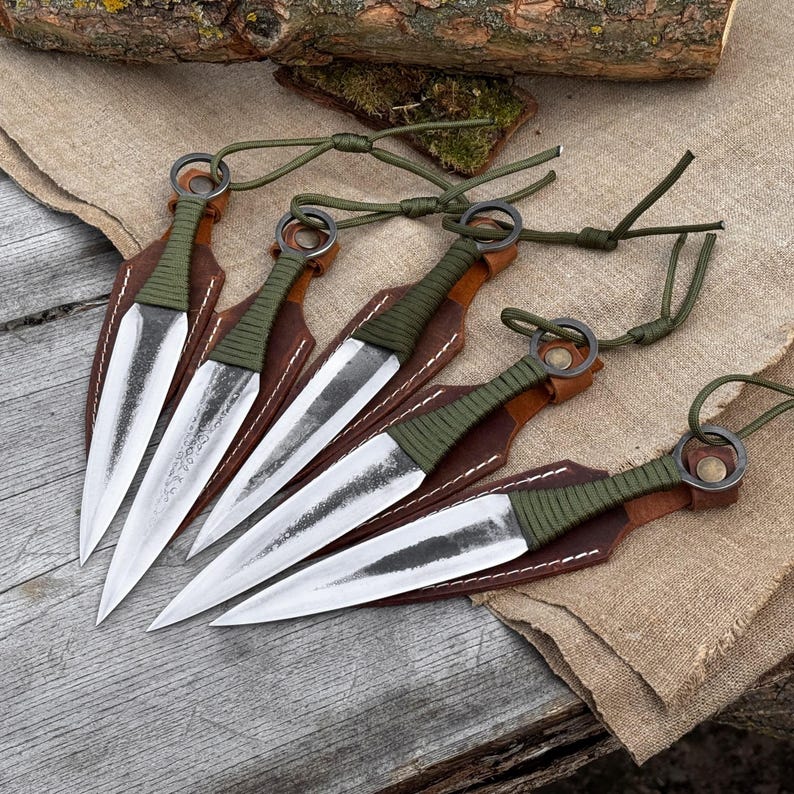 K&ouml;nnte beinhalten: F&uuml;nf Wurfmesser mit silbernen Klingen und gr&uuml;n umwickelten Griffen werden gezeigt. Jedes Messer ist in einer braunen Lederh&uuml;lle verstaut. Gr&uuml;ne Kordeln sind an den Griffen befestigt. Die Messer sind auf einer Holzoberfl&auml;che angeordnet.
