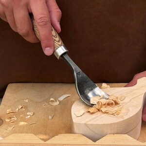 May include: A steel hook knife with a light brown wooden handle featuring a visible grain pattern. The tool is carving into a light-colored wooden block, creating small wood shavings. A small logo is stamped on the metal blade of the tool.