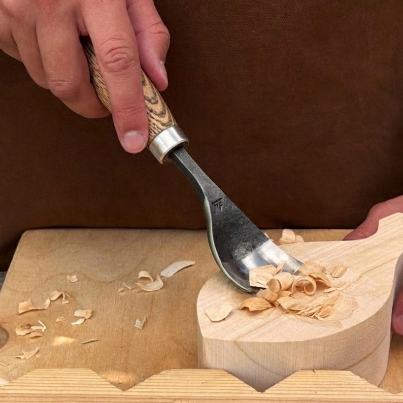 May include: A steel hook knife with a light brown wooden handle featuring a visible grain pattern. The tool is carving into a light-colored wooden block, creating small wood shavings. A small logo is stamped on the metal blade of the tool.