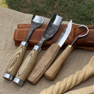 May include: A set of wood carving tools with wooden handles and metal blades. The tools include a gouge, a spoon carving knife, and a hook knife. The handles are made of light-colored wood with a natural grain pattern. A leather tool roll is in the background.