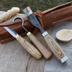 May include: A set of three wood carving tools with wooden handles and metal blades. The tools are displayed with a brown leather tool roll and a carved wooden spoon. The handles have a light wood grain pattern.