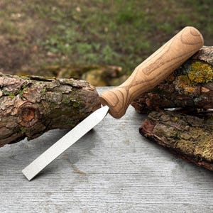Può includere: Un affilacoltelli con manico in legno e lama argentata. Il manico è in legno chiaro con un motivo a venature naturali. La lama è rettangolare e si assottiglia fino a una punta. L'affilacoltelli è appoggiato su una superficie di legno stagionato.