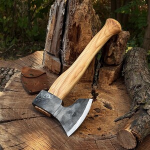 May include: A hand axe with a polished, light-coloured wooden handle and a dark, forged metal head featuring a sharp, silver-toned edge. A small, brown leather sheath with a snap closure and a stamped logo sits beside the axe head.