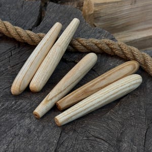 Peut inclure: Six chevilles en bois avec un motif de grain de bois naturel sont disposées sur une surface en bois sombre et texturée. Un morceau de corde est visible en arrière-plan. Les chevilles sont cylindriques et effilées.