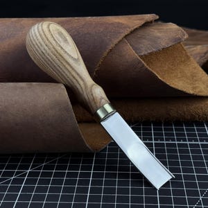Könnte beinhalten: Ein Leder-Schärfmesser mit einer polierten Metallklinge mit gerader Kante und einem abgerundeten, hellbraunen Holzgriff mit sichtbarer Maserung. Das Werkzeug verfügt über eine messingfarbene Metallzwinge, die die Klinge mit dem Griff verbindet.
