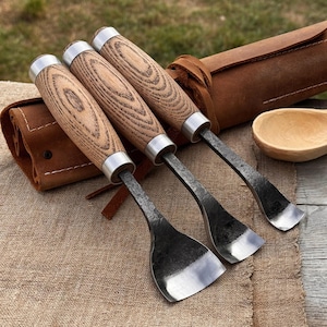 May include: Set of three wood carving chisels with wooden handles and metal blades. The chisels are displayed with a leather tool roll and a wooden spoon. The handles have a light brown wood grain pattern.