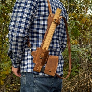 May include: A brown leather axe sheath with a shoulder strap, holding a wooden axe. The sheath has a buckle closure and a small pouch. The person is wearing a blue and white plaid shirt and blue jeans.