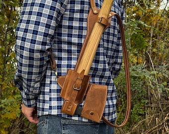 May include: A brown leather axe sheath with a shoulder strap, holding a wooden axe. The sheath has a buckle closure and a small pouch. The person is wearing a blue and white plaid shirt and blue jeans.
