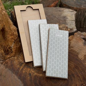 May include: A wooden sharpening stone holder with three grey sharpening stones. The stones have a white honeycomb pattern and the numbers 120, 600, and 240# printed on them. The holder is made of light-coloured wood.