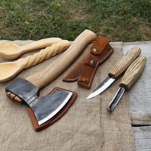Puede incluir: Un conjunto de herramientas de carpintería, que incluye un hacha con funda de cuero, dos cucharas de madera, un cuchillo con funda de cuero y dos herramientas de tallado. Las herramientas son de madera y metal, dispuestas sobre una tela beige.