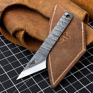 Può includere: Un coltello da marcatura in metallo grigio scuro con manico testurizzato caratterizzato da scanalature orizzontali e una lama affilata e angolata. Il coltello presenta un piccolo logo vicino al filo e un foro circolare in cima, appoggiato su un fodero in pelle marrone.