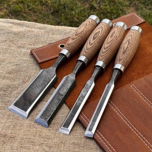 May include: A set of four wood chisels with dark, textured metal blades and light brown wooden handles. Each chisel features a silver metal ferrule connecting the blade to the handle. The tools are arranged side by side on a brown leather roll.