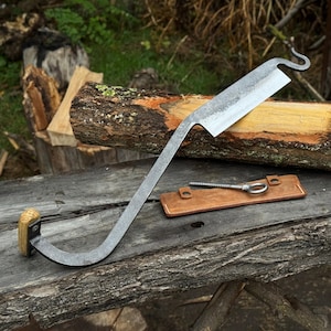 Puede incluir: Un cuchillo de tracción forjado a mano, con una hoja curva y un mango de madera. La hoja es de acero y tiene un acabado texturizado. También se incluye una funda de cuero y un tornillo de metal.