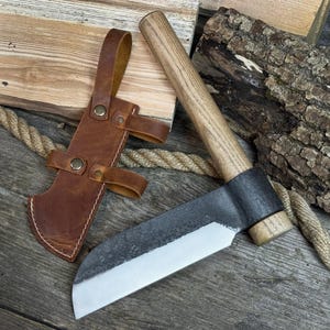 May include: A handcrafted hatchet with a wooden handle and a dark metal blade. The hatchet is accompanied by a brown leather sheath with straps. The blade is approximately 10 inches long.