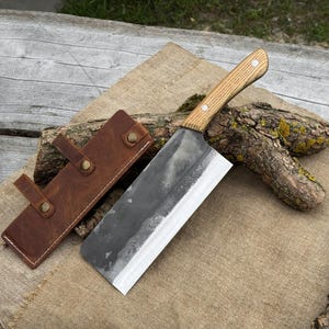 Puede incluir: Una gran cuchilla forjada a mano con un mango de madera clara y una hoja oscura y texturizada. La cuchilla descansa sobre una superficie de arpillera junto a una funda de cuero marr&oacute;n. El mango est&aacute; asegurado con dos remaches.