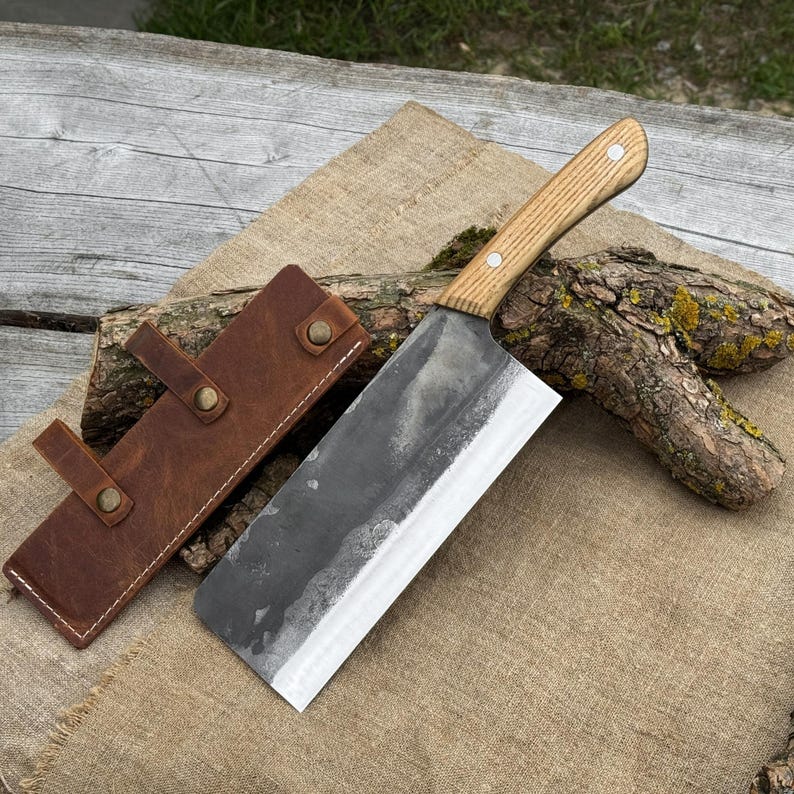 Puede incluir: Una gran cuchilla forjada a mano con un mango de madera clara y una hoja oscura y texturizada. La cuchilla descansa sobre una superficie de arpillera junto a una funda de cuero marr&oacute;n. El mango est&aacute; asegurado con dos remaches.