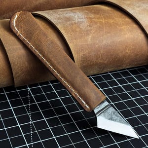 Op de afbeelding: Een hobbymes met een scherp, schuin metalen lemmet en een handvat omwikkeld met bruin leer met zichtbare witte stiksels. Het gereedschap is in een hoek geplaatst, wat het contrast tussen de textuur van het leer en het metalen lemmet benadrukt.