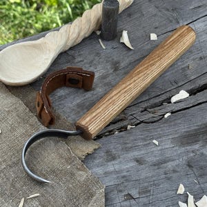 Könnte beinhalten: Ein Holzschnitzhakenmesser mit gebogener Klinge und Holzgriff. Der Griff ist aus hellbraunem Holz gefertigt. Ein Lederriemen ist am Griff befestigt. Ein geschnitzter Holzlöffel befindet sich im Hintergrund.