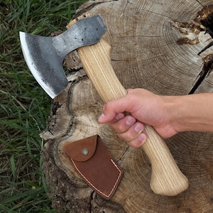 May include: A hand axe with a dark, forged steel head and a light-colored, natural wood grain handle. The axe features a curved blade edge and a sturdy grip. A small, brown leather sheath with a metal snap button is positioned near the axe head.