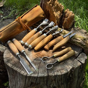 Pode incluir: Um conjunto de ferramentas de entalhe em madeira com cabos de madeira e lâminas de metal, exibidas em um tronco de árvore. O conjunto inclui cinzéis, goivas e um estojo de couro. As ferramentas têm um acabamento de madeira natural e detalhes prateados.
