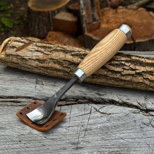 Puede incluir: Una herramienta para tallar madera con una hoja de metal oscuro curvada con una marca de fabricante visible, un mango de madera clara con virolas de metal plateado y una pequeña funda de cuero marrón cuadrada con remaches metálicos.