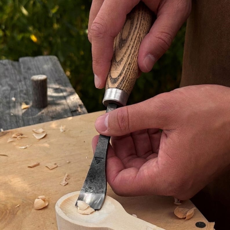 May include: A wood carving gouge with a textured wooden handle and a dark metal blade featuring a small triangular logo. The tool is being used to carve a shallow, rounded depression into a light-colored wooden spoon, creating small wood shavings.