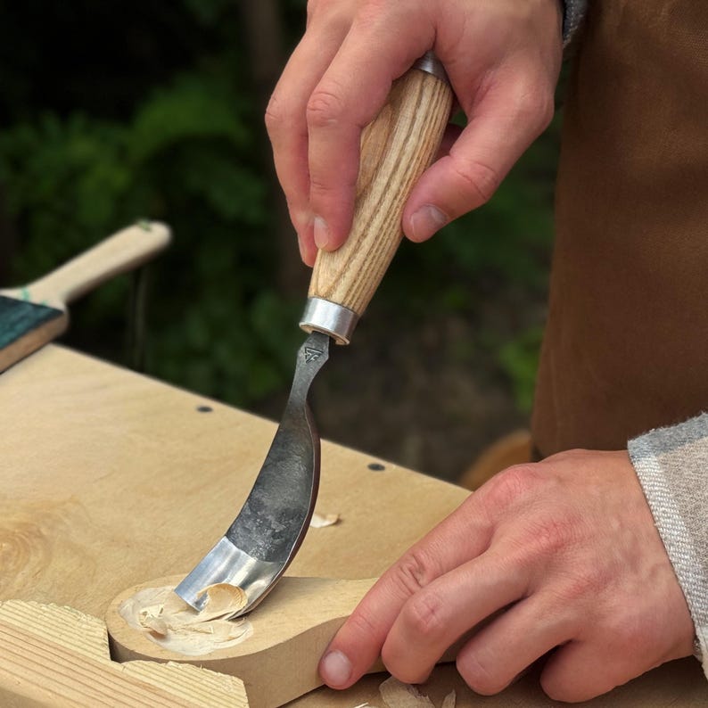 Puede incluir: Un cuchillo de gancho con una hoja de metal curva y un mango de madera clara, utilizado para tallar una cuchara de madera. La herramienta cuenta con una virola de metal plateado que conecta la hoja con el mango, con virutas de madera visibles.